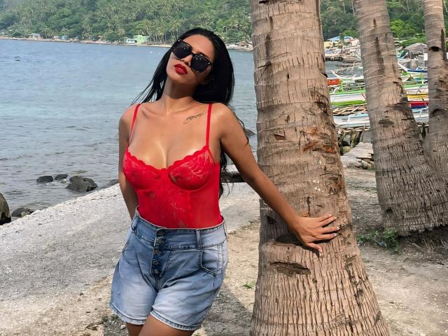 Maddison Aderson, com cabelo preto, blusa de renda vermelha, mostrando os seios, shorts jeans e óculos de sol pretos, apoiada em uma palmeira perto da água. Ela tem batom vermelho e uma tatuagem..