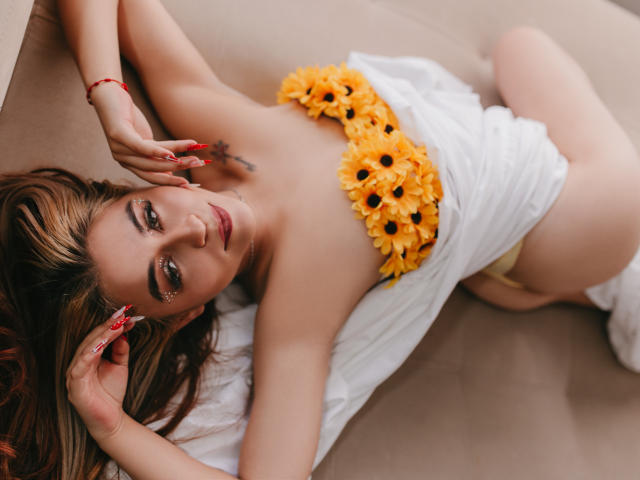 Alisses lying on her back with sunflower petals covering her tits, wearing a white cloth around her waist, and yellow panties, showing her pussy. She has red nails and a bracelet.