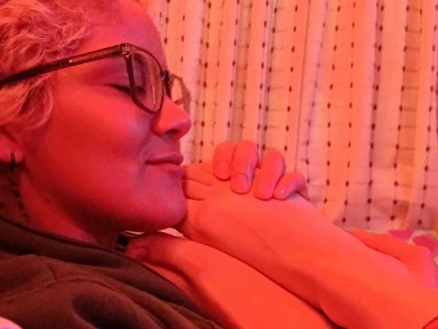 SeleneAna with curly blonde hair and glasses, holding her foot to her nose, smiling softly in red lighting, wearing a dark shirt, her toes and foot prominently in the foreground.