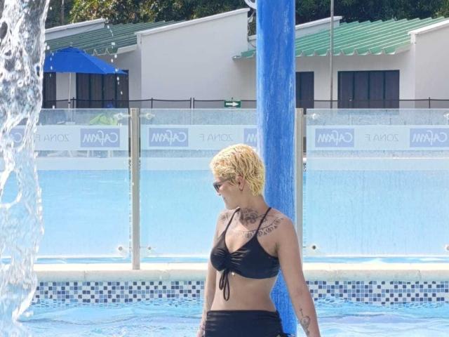 SeleneAna with short blonde hair, black bikini, tattoos on chest, standing in blue water, holding blue water column, looking down, with water splashing.
