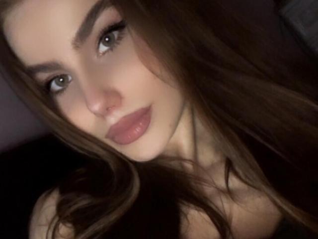 NicoleNight has fair skin, green eyes, full pink lips, and long brown hair. She's looking up with a slight smile. Her tits are not visible. The photo is dark.