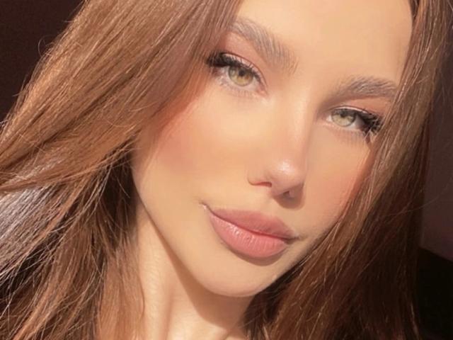 NicoleNight has brown hair, green eyes, and pink lips, with a slight smile. Her skin is smooth and sunlit, showing a natural, fresh-faced look.