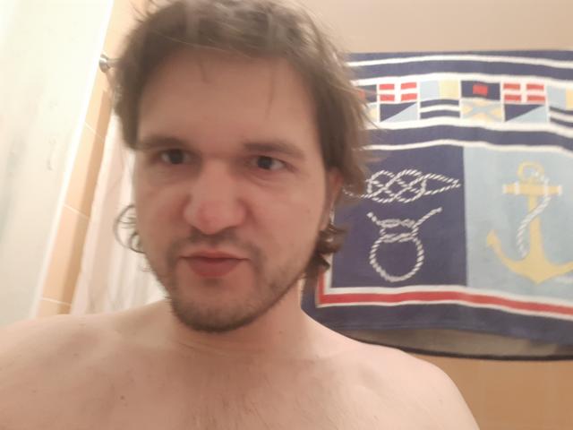 NectarineFad is a shirtless guy with brown wavy hair, a beard, and a slightly flushed face, standing in a bathroom with a navy blue anchor towel.