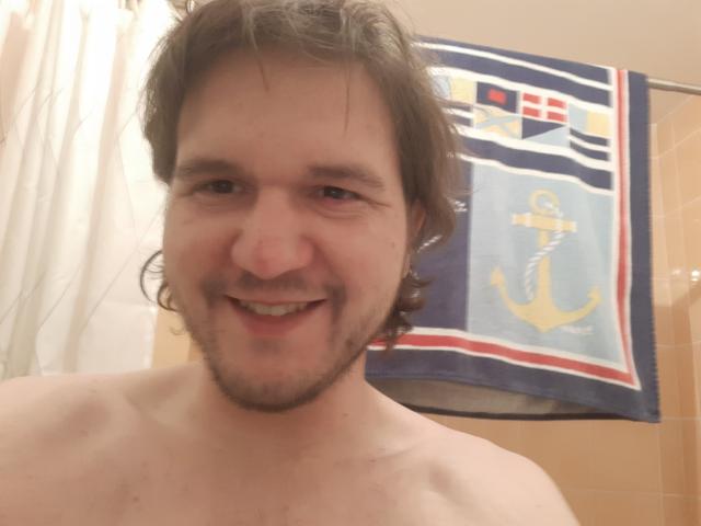 NectarineFad has a shirtless, smiling face with brown, wavy hair and a beard. His blue anchor towel with red and white stripes hangs in the background.