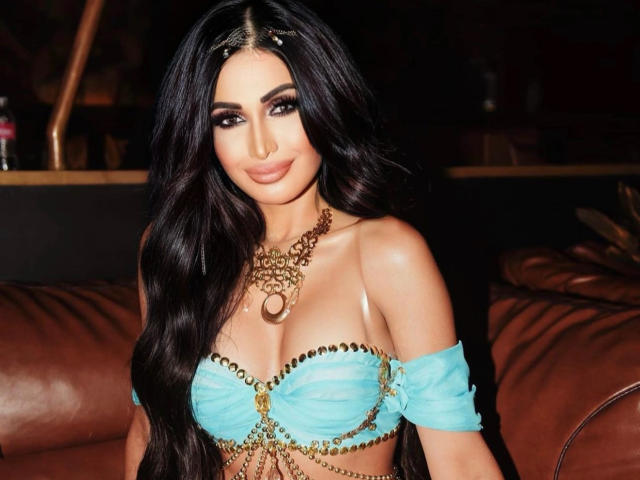 ItzMeJoanna, avec ses longs cheveux noirs, porte un haut turquoise à une épaule, orné de chaînes et de perles dorées, mettant en valeur ses seins volumineux et brillants. Elle sourit, avec un maquillage foncé et un collier en or..