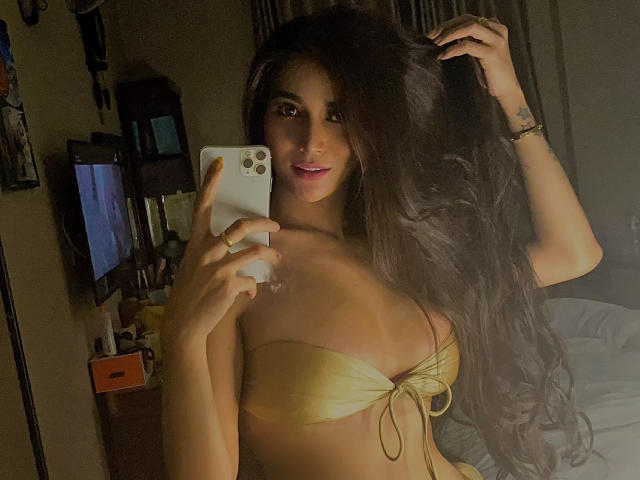 ItzMeJoanna, avec de longs cheveux noirs, un haut de maillot de bain doré, tenant un téléphone, une main dans les cheveux, souriante, montrant ses grandes seins, peau bronzée, lèvres roses, une bague au doigt, pièce faiblement éclairée..