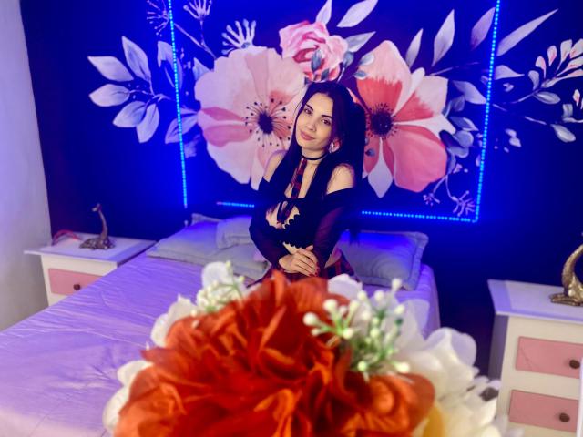 AriannaGray with long black hair, wearing a black top, sitting on a bed, arms crossed, looking at the camera, with a blue floral wall behind her and a flower in the foreground.