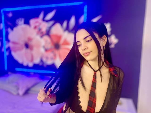 AriannaGray with long black hair, wearing a sheer black top, plaid necktie, and holding her hair, exposing her small tits. Neon blue background, floral pattern. Soft smile.