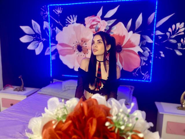 AriannaGray with long black hair, wearing a black top with red and black straps, sitting on a bed, blue LED floral wall art, and white floral decor in foreground.