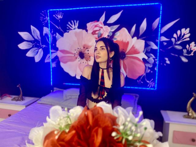 AriannaGray with long black hair, wearing a black off-shoulder top, looking up, has a cute face, small tits, and a slim figure, in a bedroom with floral LED lights.