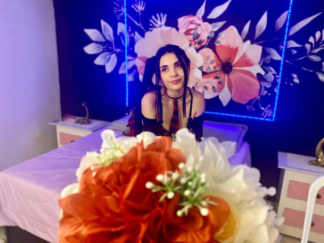 AriannaGray with long dark hair, wearing a black off-shoulder top, sits on a bed, looking at the camera, with colorful flowers in the foreground, purple blue LED lights.