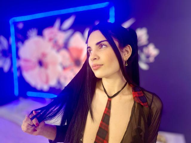 AriannaGray with long black hair, wearing a sheer black top, red plaid tie, and earrings, holding her hair, smiling, purple blue neon background, floral screen.