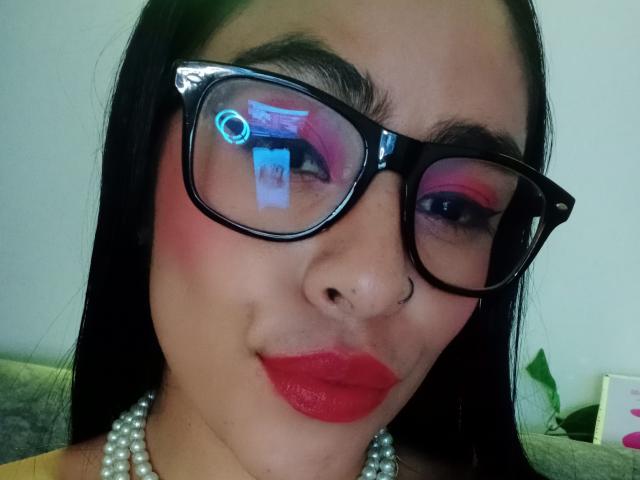 StellaBeckker con occhiali neri, rossetto rosso, ombretto rosa, piercing al naso, collana di perle e riflesso di luce blu negli occhiali, che mostra un leggero sorriso..