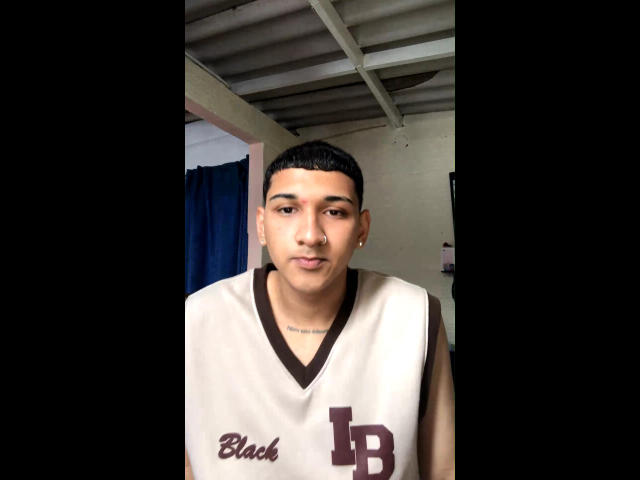 Dekk, short black-haired dude, wearing a sleeveless white basketball jersey with brown letters, small tattoo on chest, standing in doorway, looking slightly to the side, neutral expression.