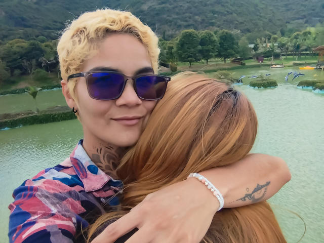 SeleneAna, mit blonden Locken, blauen Sonnenbrillen und einem pinken Karohemd, umarmt eine Frau mit langen braunen Haaren, einem weißen Armband und einem Tattoo auf ihrem Arm und lächelt in die Kamera..