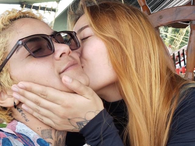 SeleneAna with blonde hair and glasses is kissing the cheek of a man with curly hair and tattoos. Her hand is on his face. She's wearing a black shirt.