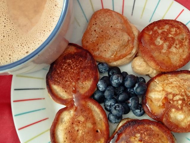 A plate of golden-brown pancakes, crispy on the edges, with a side of fresh blueberries and a cup of frothy, beige coffee. SeleneAna's breakfast.