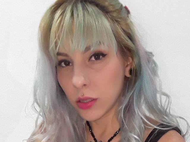 Chloe69Blauw, met pastelblauw en blond haar, pony, zwarte eyeliner, neusring, rode lippen, een zwarte kralenketting en kleine zwarte oorbelgaatjes. Lichte huid, zachte uitdrukking..