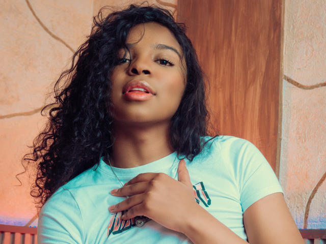 LisaMendez with curly black hair, light green shirt, and pink lipstick, tongue slightly out, hand touching chest, looking at camera with a subtle smile.
