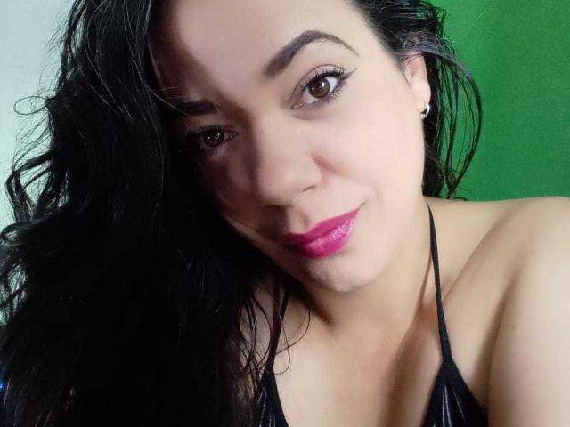 YolittS has black wavy hair, brown eyes, pink lipstick, small hoop earrings, and a black halter top showing her tits. She's smiling subtly with a green background.