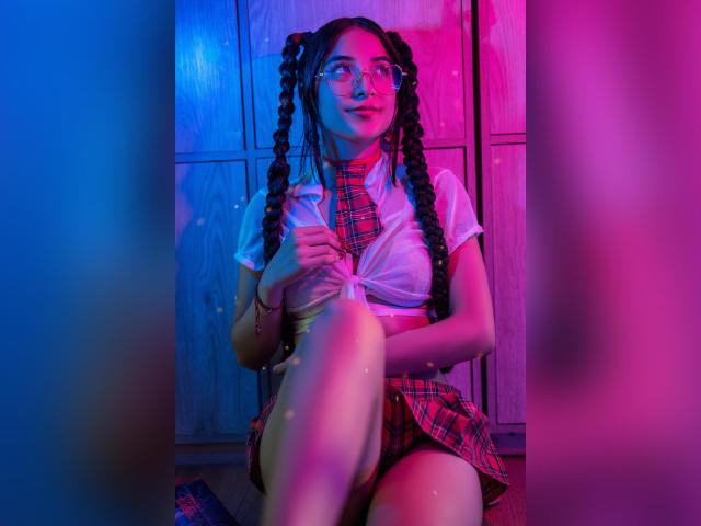 ValeriaClark with glasses, tied-up pigtails, white tied shirt, plaid skirt, and red tie, sitting, legs crossed, under blue and pink neon lights, smiling, showing her tits.