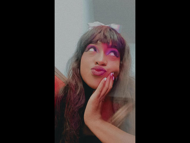 LilaJamez with long brown hair, pink eye makeup, white bow, pouty lips, hand on cheek, pink nail polish, looking up, soft light, slightly blurred, cute, sexy.