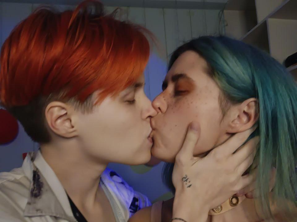 CrystalFemmes with red and blue hair kiss, eyes closed. Red-haired CrystalFemmes holds blue-haired CrystalFemmes' face, finger tattoo visible. Soft light, intimate moment.