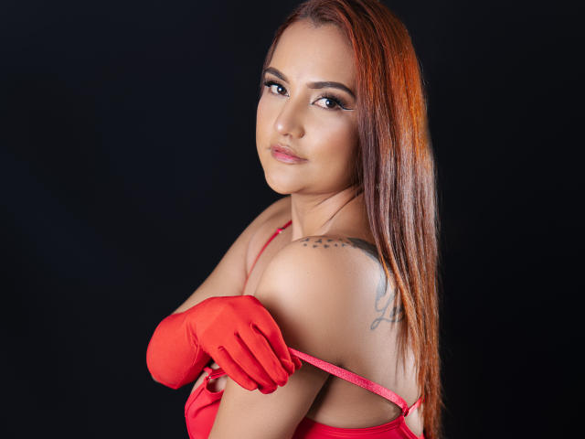 SamyPrinces with red gloves, red dress, long brown hair, tattoo on right shoulder, looking over shoulder, black background, slight smile, sexy, attractive, red lipstick, bare shoulders.