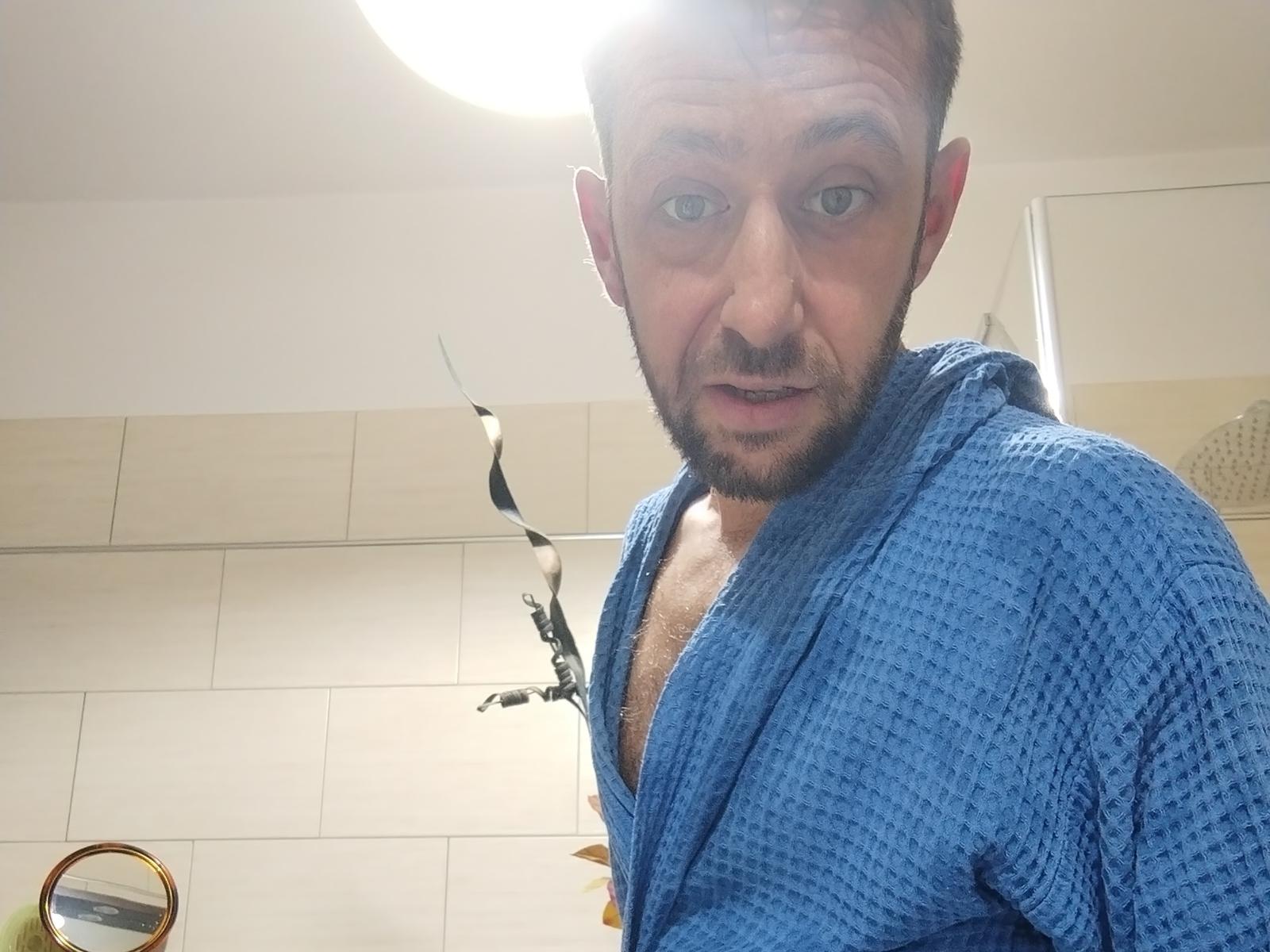 SteuHot with a blue waffle-texture bathrobe, partially open to reveal his hairy chest, looking at the camera with a slightly surprised expression. Light from above.