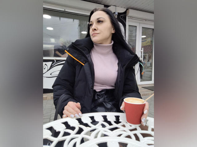XCrazyBabeX with dark hair, black coat, pink turtleneck, holding red cup, white metal table, looking to the side, gray background, overcast day, slight frown, white nails.