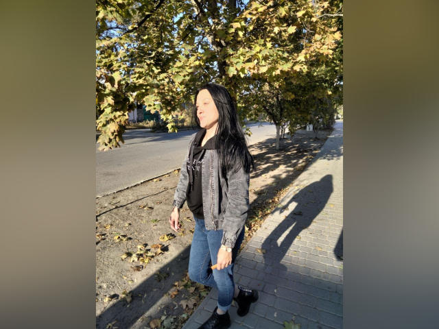 XCrazyBabeX with long black hair, gray jacket, black shirt, blue jeans, and black shoes, standing on a sunlit sidewalk, shadow on the ground, autumn leaves in the background.