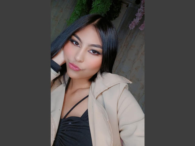 CarlaRusso with black hair, tan coat, and black top, has pink lips, dark eyeliner, and a cute smile. She's looking at the camera with a slight tilt.