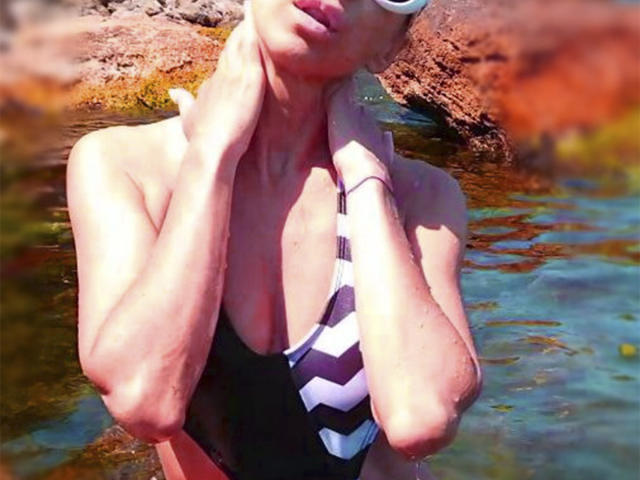 KatarinaDamo in a black and white striped one-piece swimsuit, standing in shallow water with hands near her neck, showing her tits and a slight tan.