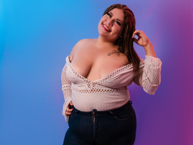 CandyFuckBoobs with big tits, wearing a white off-shoulder top and dark jeans, smiling, hand in hair, tattoo on right shoulder, blue and purple gradient background.
