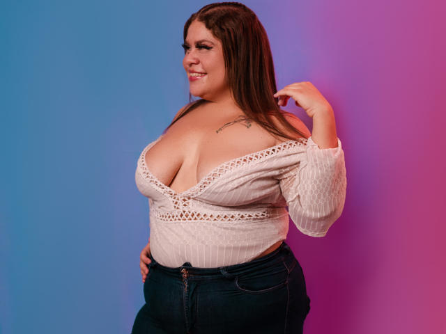 CandyFuckBoobs with large tits, wearing a white, long-sleeve, lace-up top, dark jeans, and long brown hair, smiling and touching her hair. She has a tattoo on her chest.