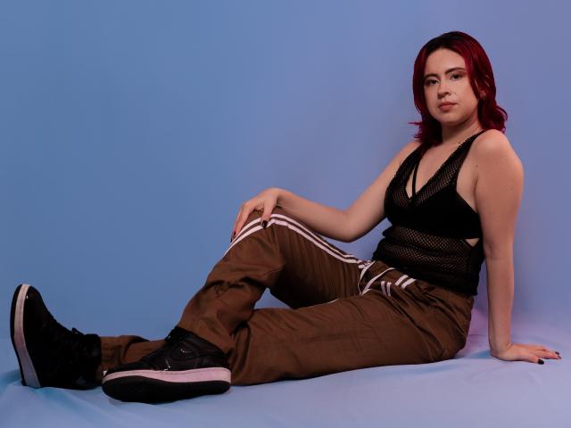 NatySilvy with red hair, black mesh top, brown pants, and black sneakers, sitting on the floor, showing her ass and small tits, against a blue background.