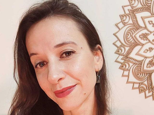 BrendaSun has light skin, brown eyes, and long dark hair. She's smiling with red lipstick, wearing small earrings, and has a small mole on her right cheek.