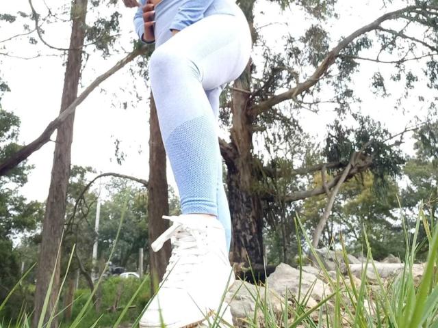 Victoria Waynes con i suoi glutei in vista, indossa dei leggings azzurri chiari, aderenti e con motivo a rete, è in piedi con una gamba sollevata, mostrando delle scarpe da ginnastica bianche con lacci, in una zona erbosa e boscosa..