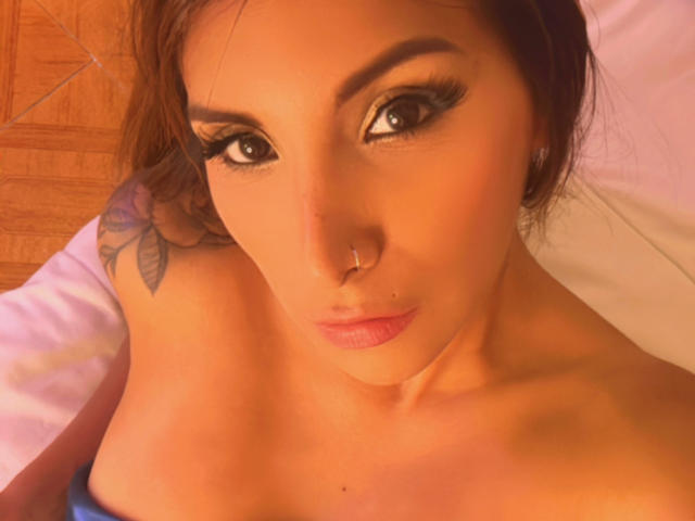Stephaniez with big, dark eyes, heavy eyeliner, and pink lips, looking up at the camera. She has a nose ring, tattoo on her right shoulder, and exposed boobs.