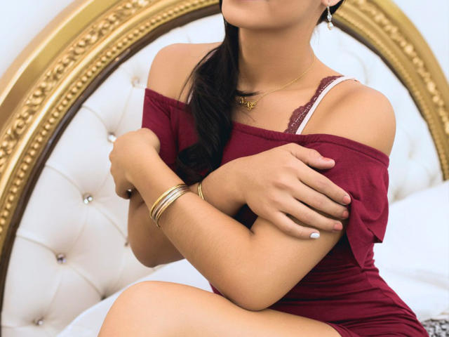 AlessiaMistery with her arms crossed, wearing a red off-shoulder top, showing her small tits, white nail polish, and gold bracelets, sitting on a bed.