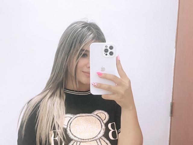 SashaPolet with blonde hair, smiling, holding an iPhone, wearing a black bear shirt, pink nails, white wall, and pink door in the background. Boobs not visible.