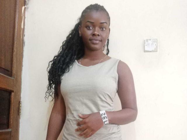 Beautyqu with dark skin, long curly black hair, wearing a beige sleeveless top, standing, right hand on belly, white wristband with black letters, looking at camera, neutral expression.