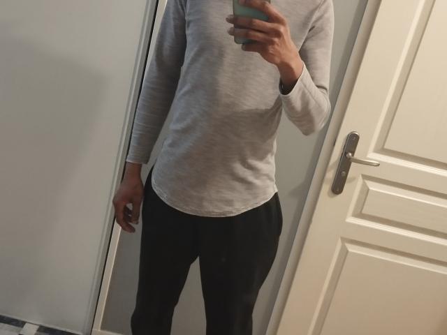 ChaChaHott in a grey long-sleeve shirt and black pants, holding a phone in front of a mirror, with a door and door handle in the background.