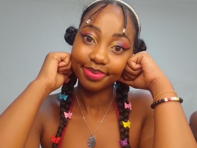 KendyQ with dark skin, pink lipstick, colorful braids, butterfly clips, pearl necklace, and pink eyeshadow, smiling, hands near ears, wearing bead bracelet, pearl headband.