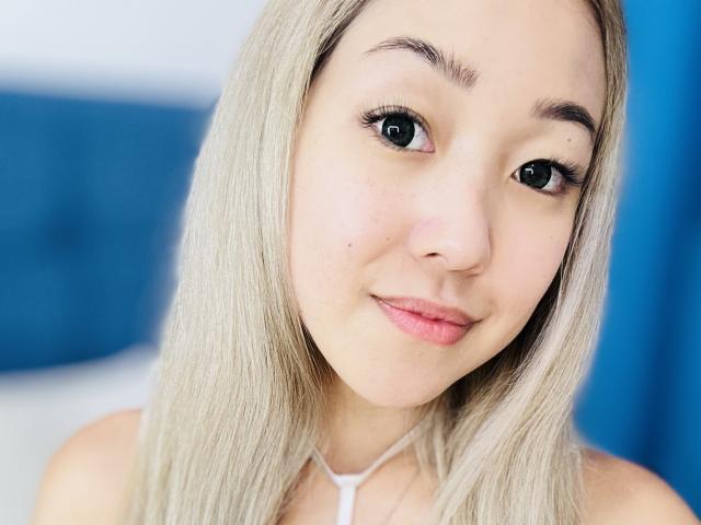 RenyLime has blonde hair, green eyes, and a small nose. She's smiling with pink lips, wearing a white necklace, and has a light skin tone. Her tits are not visible.