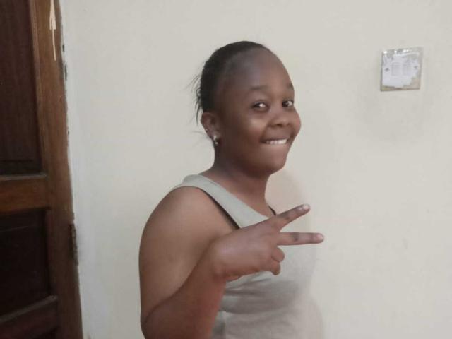 Bootyque with dark skin, smiling, pointing with right index finger, wearing a gray tank top, small earrings, and hair in a bun.