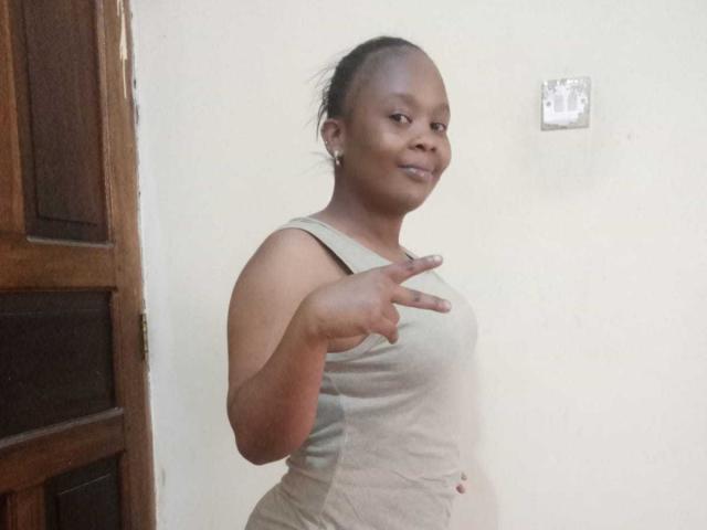 Bootyque with short hair, medium brown skin, small breasts, wearing a beige tank top, pointing forward, smiling slightly, standing next to a wooden door.