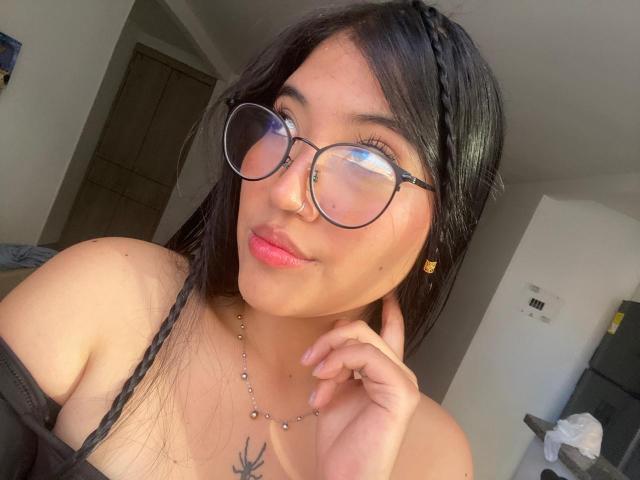 Elizaa mit runder Brille, Nasenring, geflochtenem schwarzen Haar, rosafarbenen Lippen, einer Halskette und einem Brusttattoo, die ihre Kehle berührt, wobei ihre nackten Schultern und kleinen Brüste zu sehen sind..