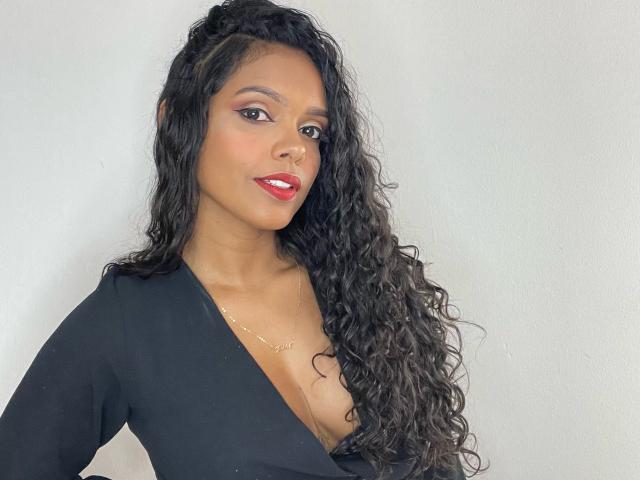 CarollyneRms with long curly black hair, red lipstick, and black deep V-neck top, showing her tits, looking at camera, light skin, small gold necklace.