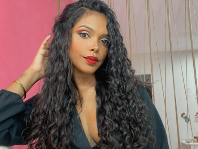 CarollyneRms with long curly black hair, red lipstick, and black top, hand in hair, looking at camera. Her tits are covered, but her cleavage is visible.